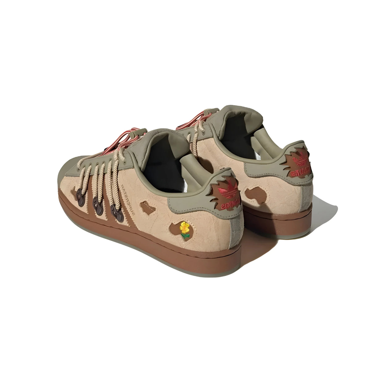 A pair of adidas melting sadness superstars sneakers shown from a three-quarters rear angle on a white background. The shoes feature a beige canvas upper with olive green suede overlays, brown soles, and small embroidered patches including a red maple leaf and a yellow/orange bird.