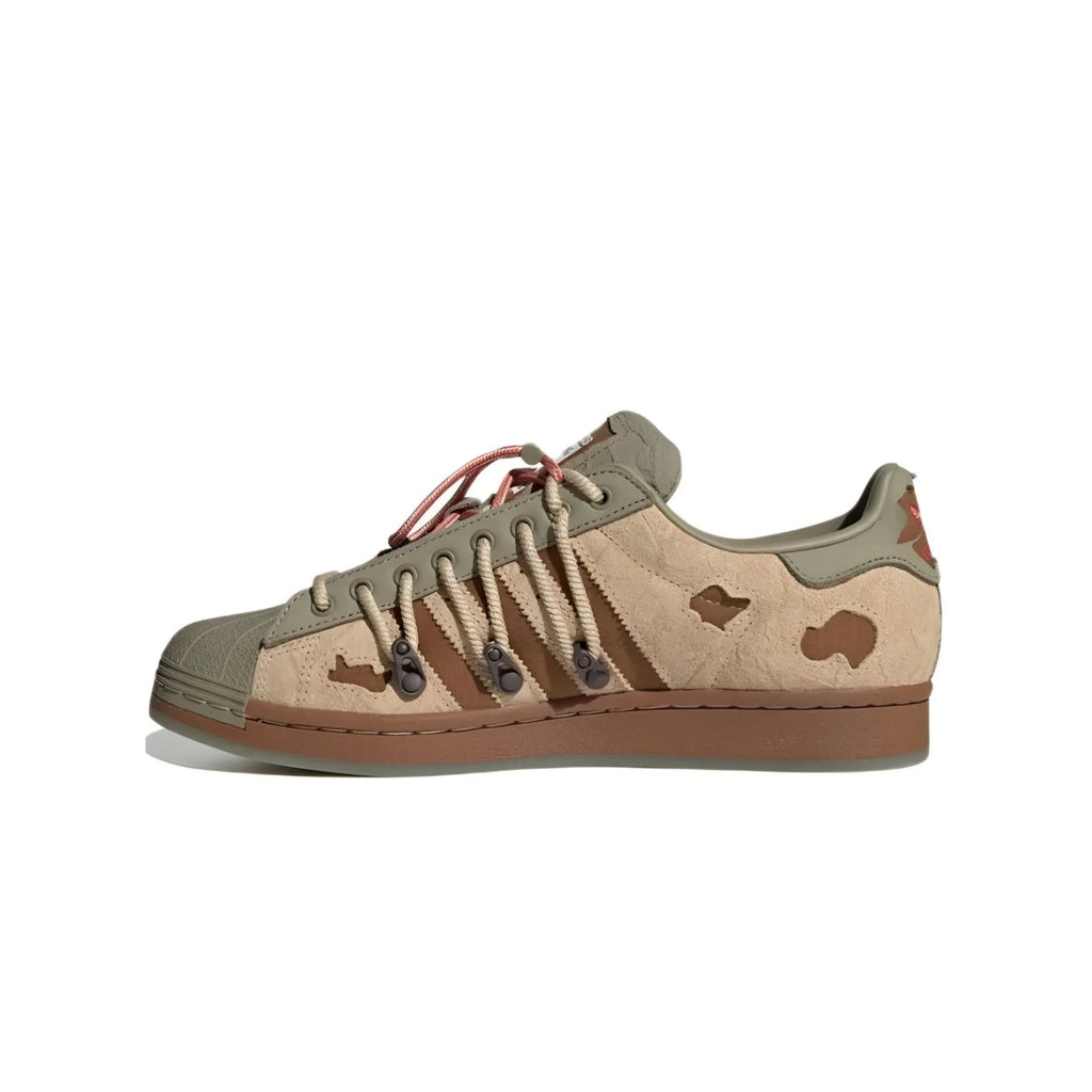 Adidas superstar in a muted camouflage pattern of olive green, tan, and brown, featuring a gum sole and matching brown laces with dark metal aglets.