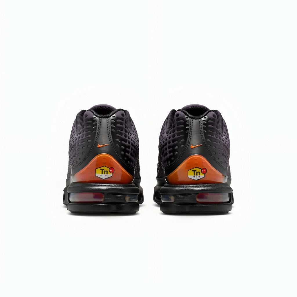 A pair of Nike Air Max plus VII sneakers shown from the back, featuring a black and dark gray textured upper, a bright orange heel accent with a yellow 'in' logo, and a black sole with a visible air cushion unit.